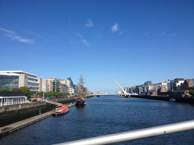 In this image I can see few building,trees,ships,water and bridge. The sky is in blue and white...