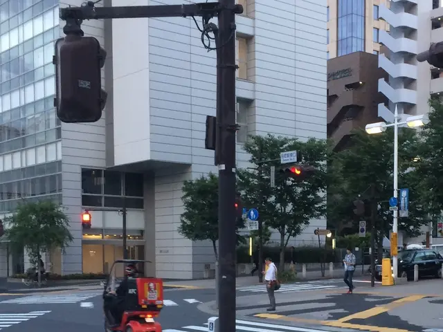 In the image there is a scooter on the road and few persons standing in the back, in the front...