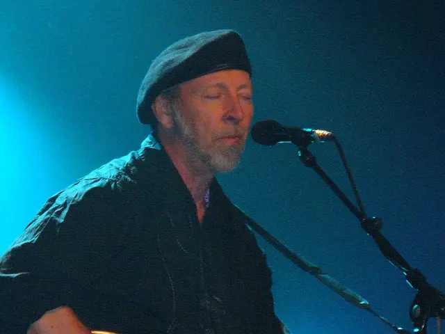 In this picture we can see a man in balck attire and wearing a black cap, standing in front of mike...