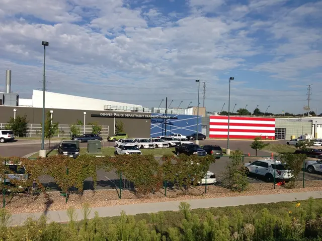 In this image we can see cars, trees, road, building, poles, sky and clouds.