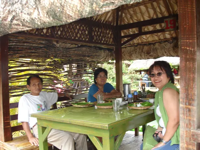 Under this tent these three persons are sitting on a bench. On this table there are plates, glasses...